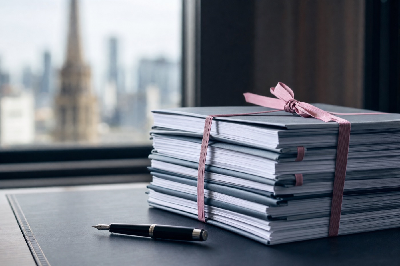 A stack of bound court pleadings tied with pink legal ribbon beside a fountain pen on a dark chambers desk in Melbourne